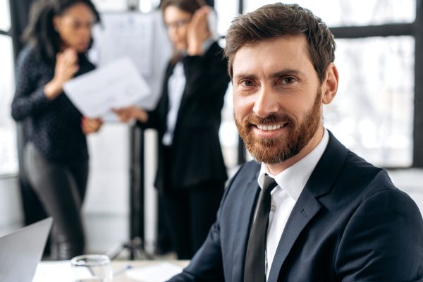 Confident elegant business man, company ceo, sits in office during corporate brainstorm, looks at camera, smiles. Portrait of attractive caucasian male business leader in a suit at briefing meeting