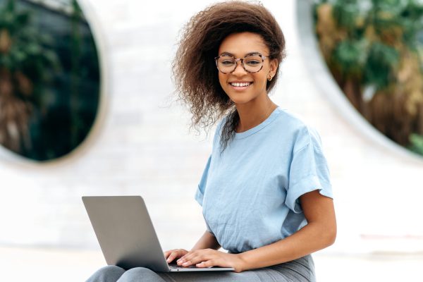 Photo of an elegant lovely positive confident African American woman, with glasses, manager, company seo, freelancer, sits with laptop outdoors near business center, looks at camera, smiles friendly