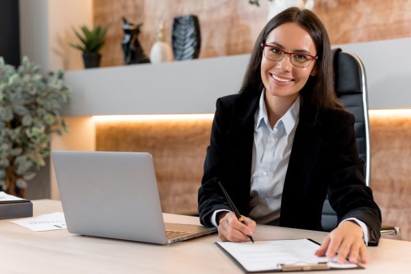 Portrait of a charming, intelligent Caucasian woman with glasses, office worker, a management company, trader investor, sit at work desk in modern office, in stylish business clothes, smiling